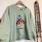 A sage green crewneck sweatshirt hanging on a wooden hanger against a textured white wall. The sweatshirt features a graphic print of a cowboy riding a bucking brown horse on the front.