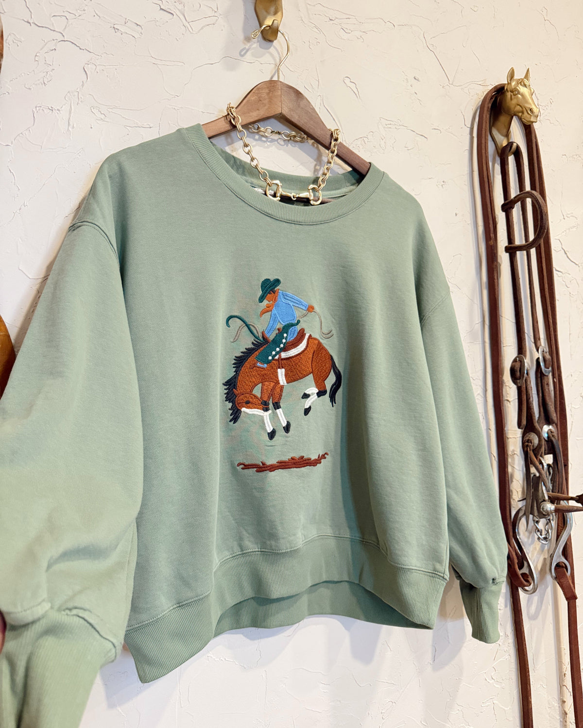 A sage green crewneck sweatshirt hanging on a wooden hanger against a textured white wall. The sweatshirt features a graphic print of a cowboy riding a bucking brown horse on the front.