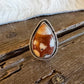 A teardrop-shaped silver ring with a carved brown and cream stone, resting on a rustic wooden surface next to a white fur trim.