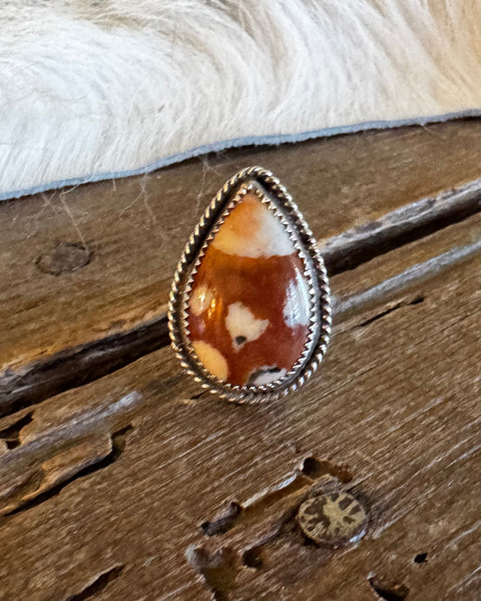 A teardrop-shaped silver ring with a carved brown and cream stone, resting on a rustic wooden surface next to a white fur trim.