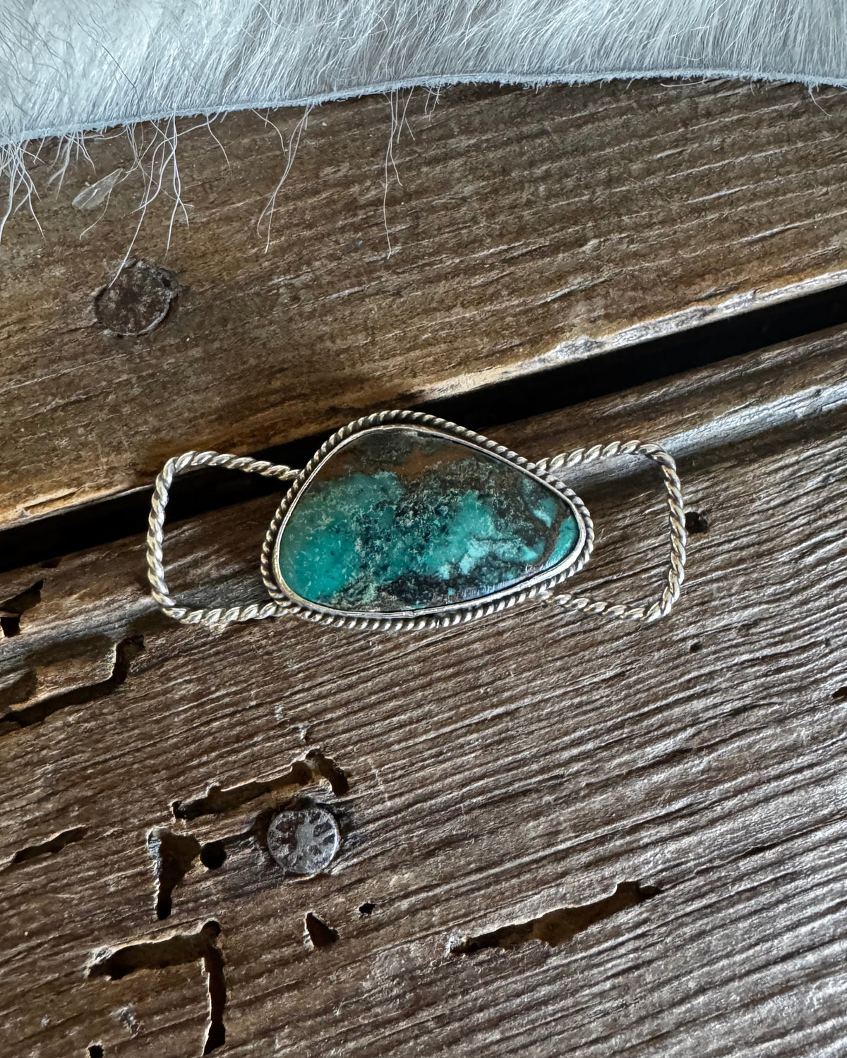 A Southwestern-style sterling silver ring featuring a large, irregular turquoise stone with natural brown and black matrix inclusions, resting on a rustic weathered wooden surface.