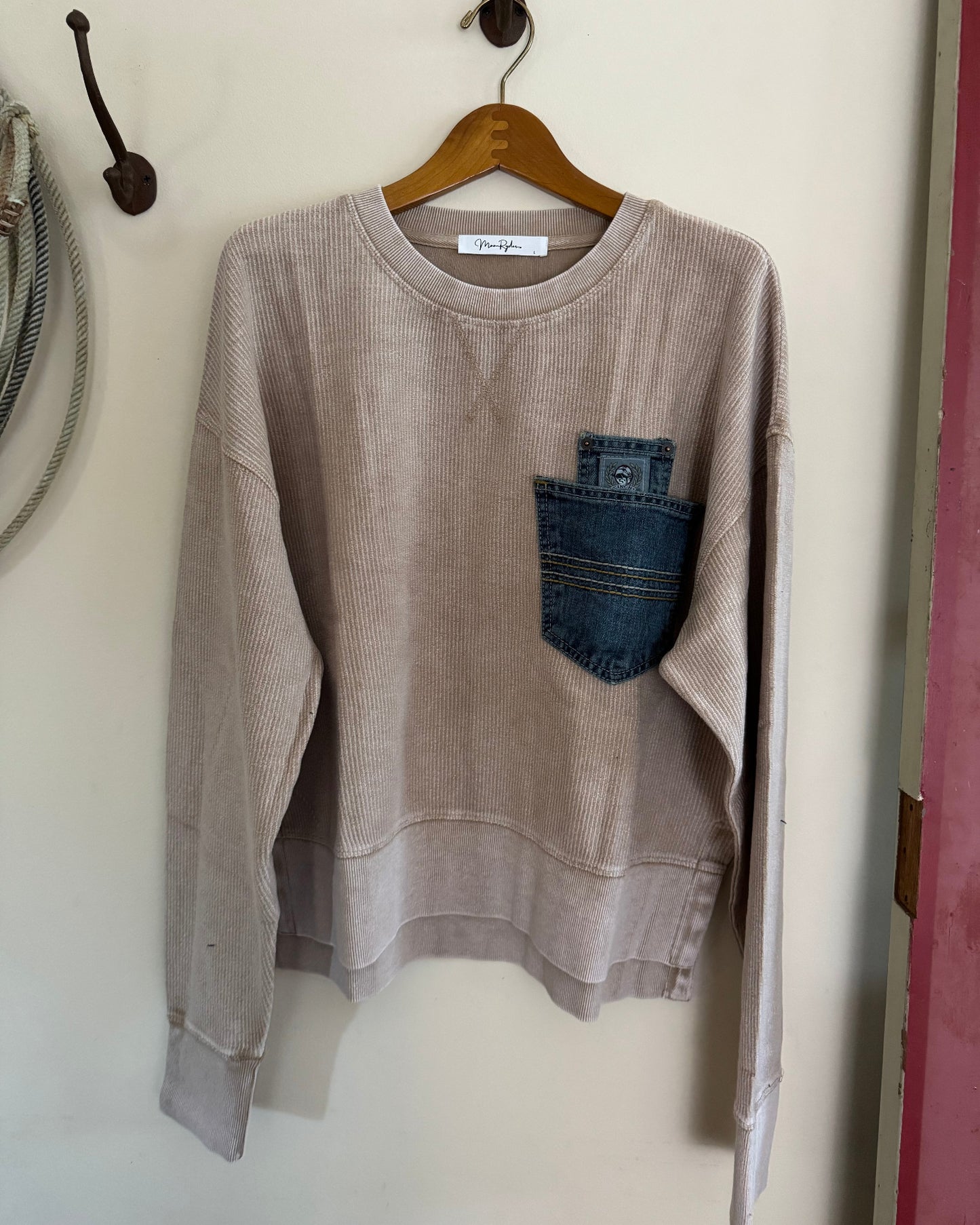 Brown sweatshirt with a denim pocket on a hanger against a white wall.