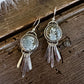 A pair of handmade silver-toned dangle earrings featuring round bezel-set gray jasper stones and hanging clear quartz crystal strands, displayed on a rustic dark wood surface with a white faux fur accent.
