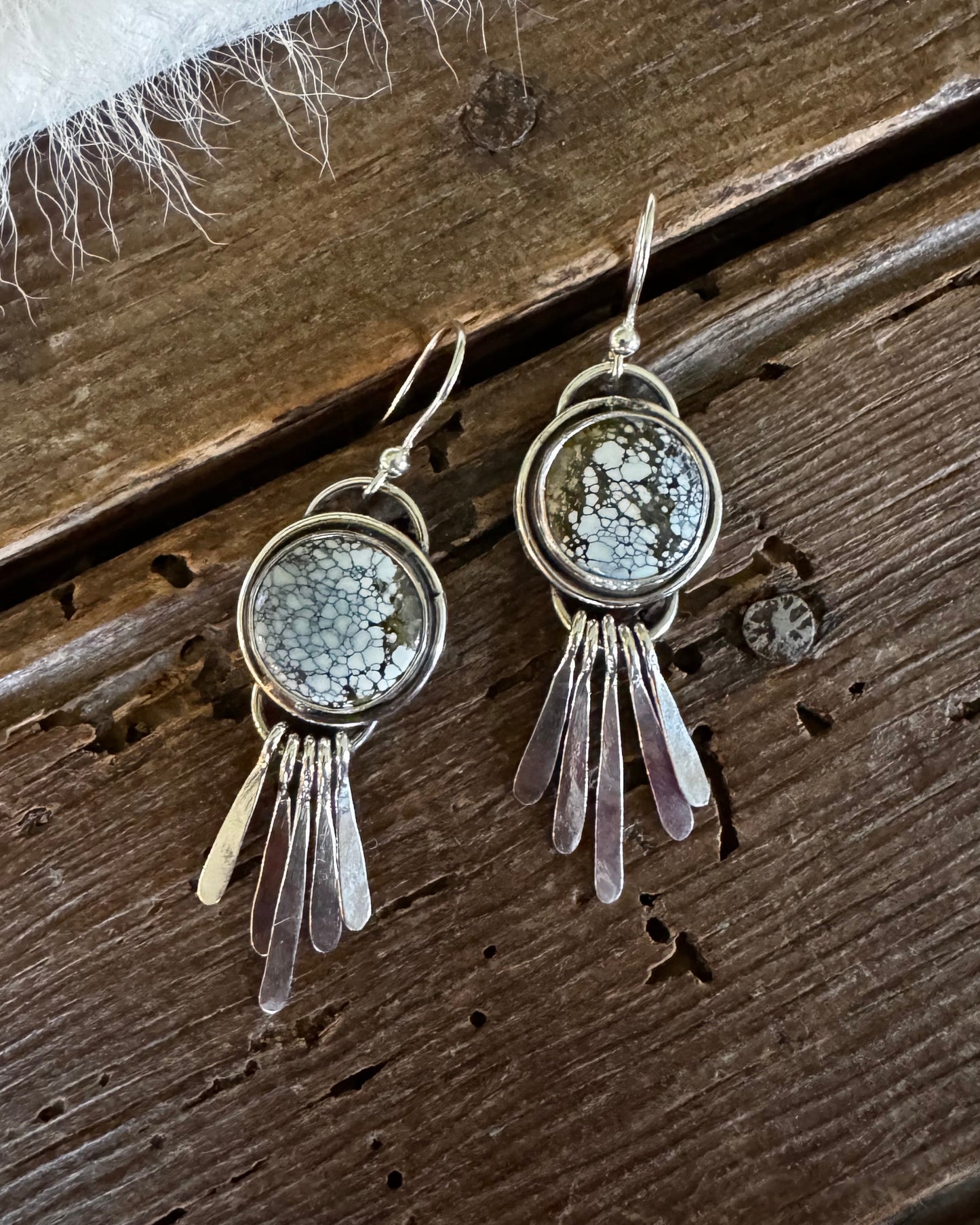 A pair of handmade silver-toned dangle earrings featuring round bezel-set gray jasper stones and hanging clear quartz crystal strands, displayed on a rustic dark wood surface with a white faux fur accent.