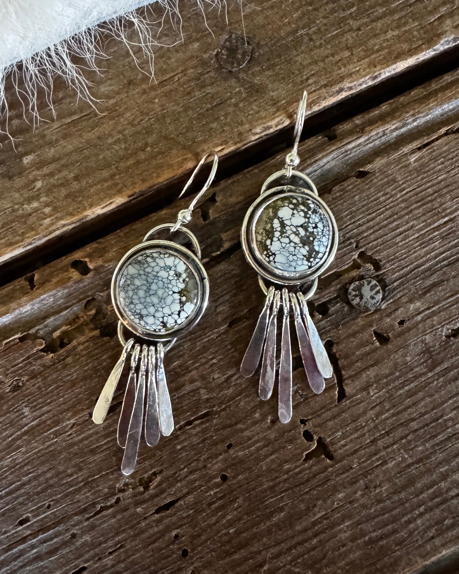 A pair of handmade silver-toned dangle earrings featuring round bezel-set gray jasper stones and hanging clear quartz crystal strands, displayed on a rustic dark wood surface with a white faux fur accent.