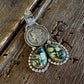 A silver-toned pendant necklace featuring a circular coin medallion and two dangling teardrop-shaped turquoise-like stones with green, blue, and brown matrix, displayed on a rustic dark wood surface.