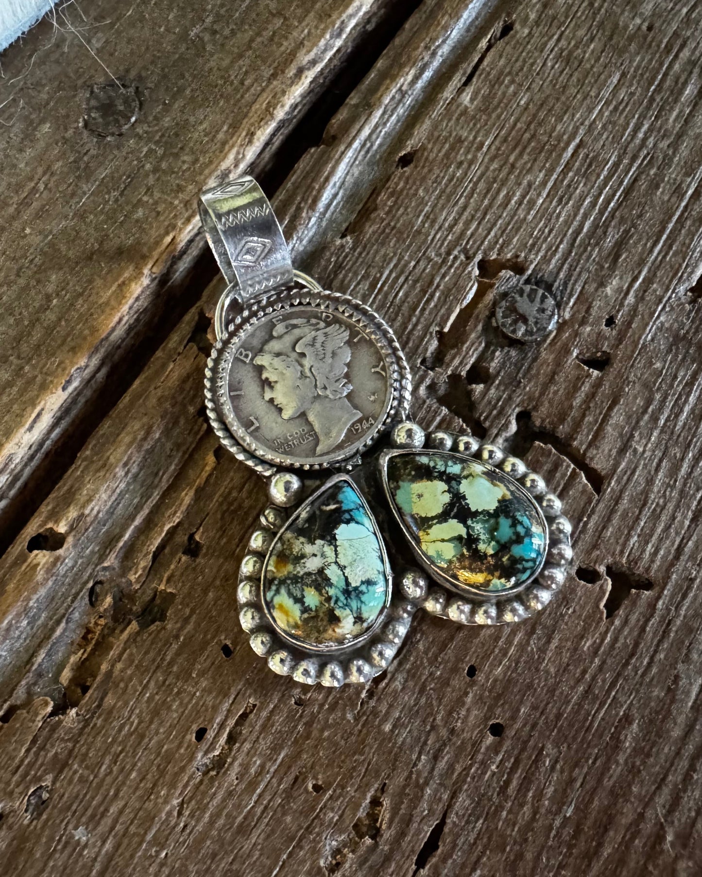 A silver-toned pendant necklace featuring a circular coin medallion and two dangling teardrop-shaped turquoise-like stones with green, blue, and brown matrix, displayed on a rustic dark wood surface.