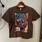 A brown short-sleeve t-shirt hanging on a wooden hanger against a plain off-white wall. The shirt features a large rectangular graphic print on the front with an abstract painting depicting a cowboy boot and rope.