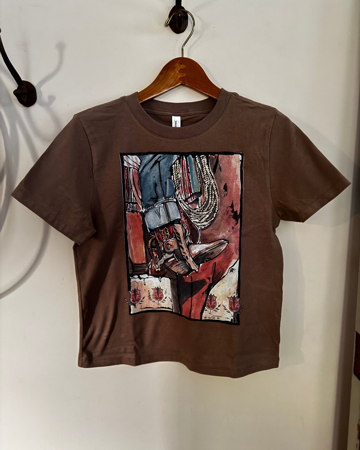 A brown short-sleeve t-shirt hanging on a wooden hanger against a plain off-white wall. The shirt features a large rectangular graphic print on the front with an abstract painting depicting a cowboy boot and rope.