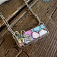 A rectangular pendant necklace with a colorful mosaic pattern of pink, blue, and white pieces, set in a silver-toned frame and hanging from a silver chain on a rustic wooden background.