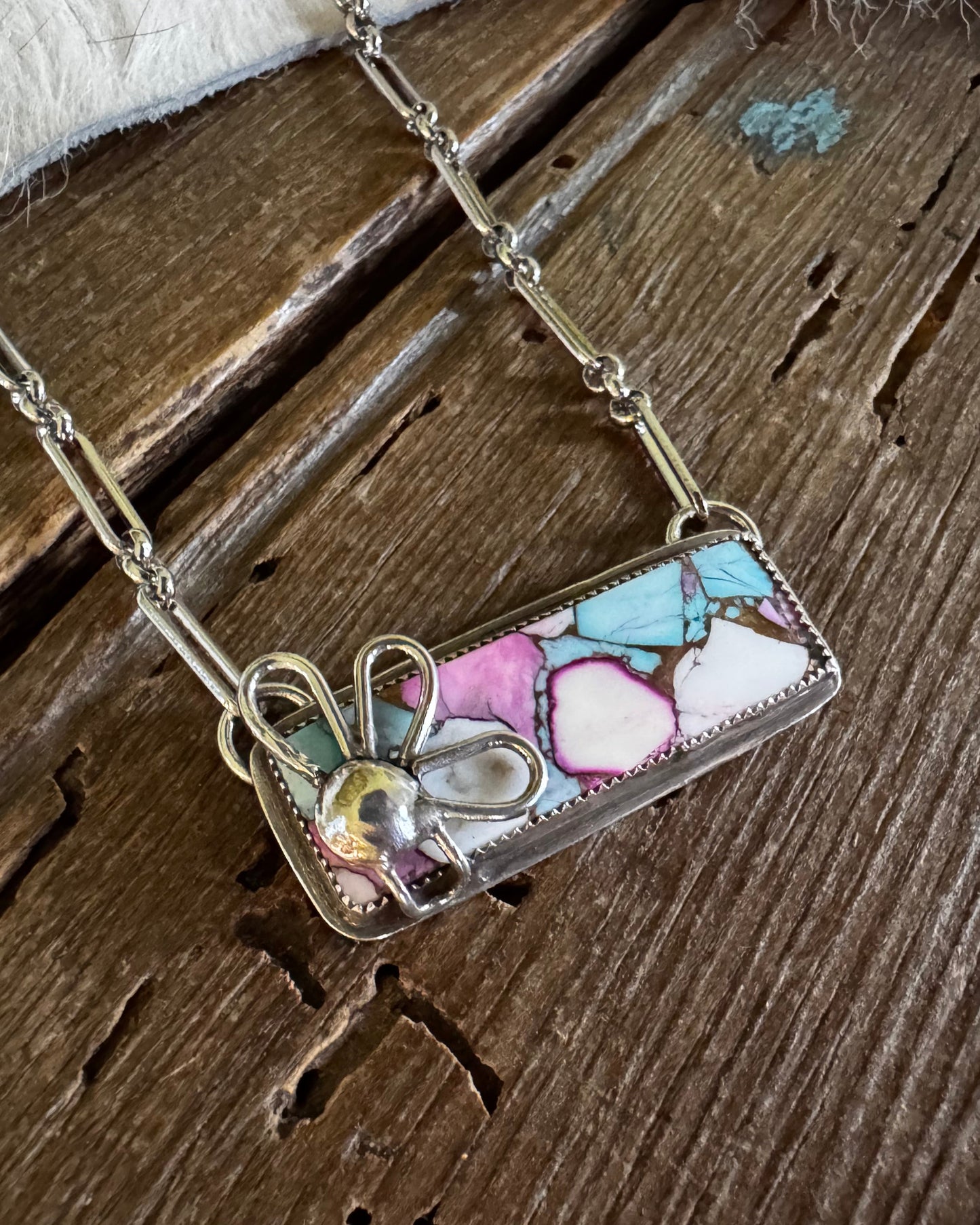 A rectangular pendant necklace with a colorful mosaic pattern of pink, blue, and white pieces, set in a silver-toned frame and hanging from a silver chain on a rustic wooden background.