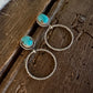 A pair of gold-toned drop earrings with oval turquoise-blue stones, displayed on a rustic dark wood surface next to a white fur trim.