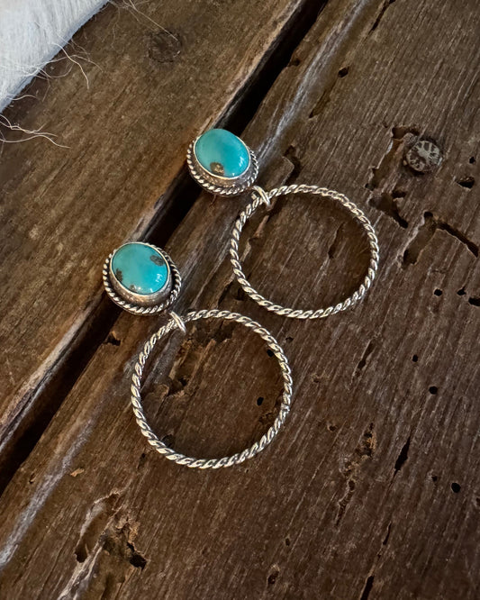 A pair of gold-toned drop earrings with oval turquoise-blue stones, displayed on a rustic dark wood surface next to a white fur trim.