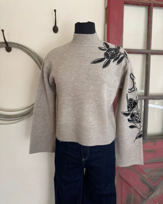 Gray sweater with black floral embroidery on a mannequin against a neutral wall.