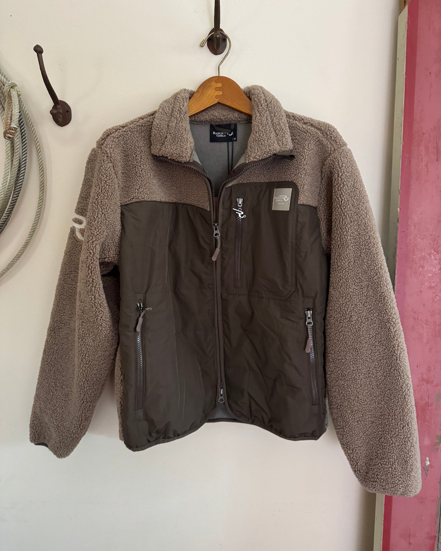 Two-tone jacket with brown fleece collar and black body on a wooden hanger against a white wall.