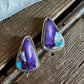 A pair of triangular stud earrings with purple and blue agate inlays set in silver, resting on a rustic wooden surface next to a white faux fur trim.