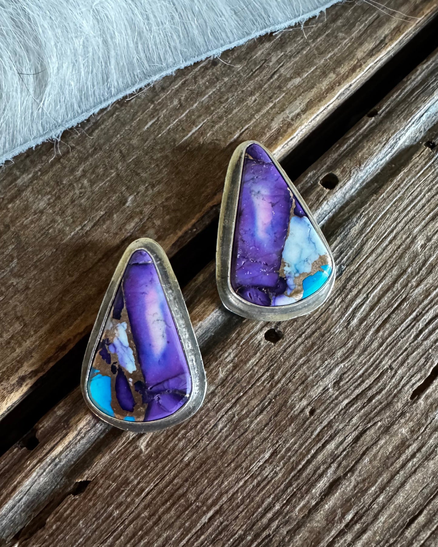 A pair of triangular stud earrings with purple and blue agate inlays set in silver, resting on a rustic wooden surface next to a white faux fur trim.