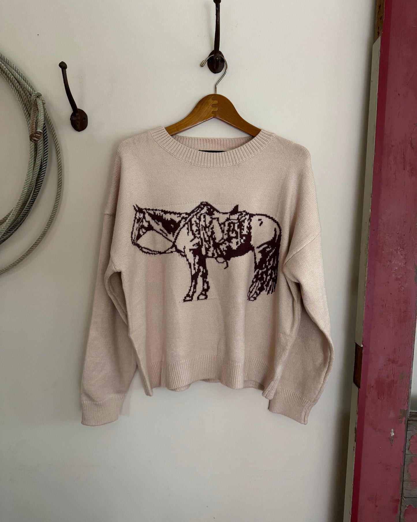 Beige sweater with a brown graphic design on a wooden hanger against a white wall.