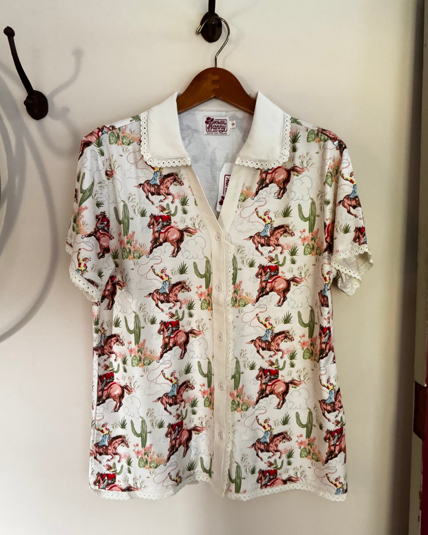 A short-sleeve button-up shirt with a white base and a colorful cowboy print, hanging on a wooden hanger against a plain wall.