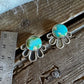 A pair of silver-toned daisy-style earrings with round turquoise-blue cabochon stones, displayed on a rustic wooden surface next to a metal ruler for scale.