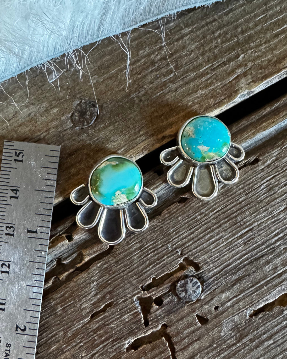A pair of silver-toned daisy-style earrings with round turquoise-blue cabochon stones, displayed on a rustic wooden surface next to a metal ruler for scale.