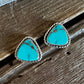 A pair of triangular turquoise stud earrings with silver rope borders, resting on a rustic dark wood surface with a white faux fur accent at the top.