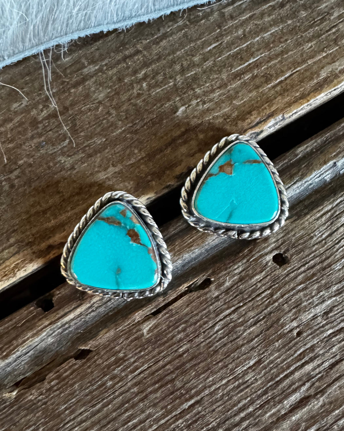 A pair of triangular turquoise stud earrings with silver rope borders, resting on a rustic dark wood surface with a white faux fur accent at the top.