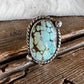 A silver ring featuring an oval turquoise stone with brown matrix veining, displayed on a rustic wooden surface with a white faux fur accent.