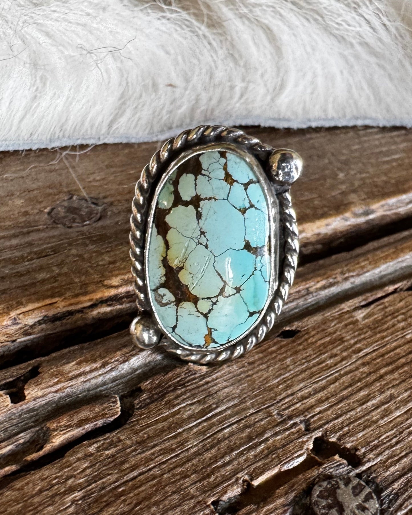 A silver ring featuring an oval turquoise stone with brown matrix veining, displayed on a rustic wooden surface with a white faux fur accent.