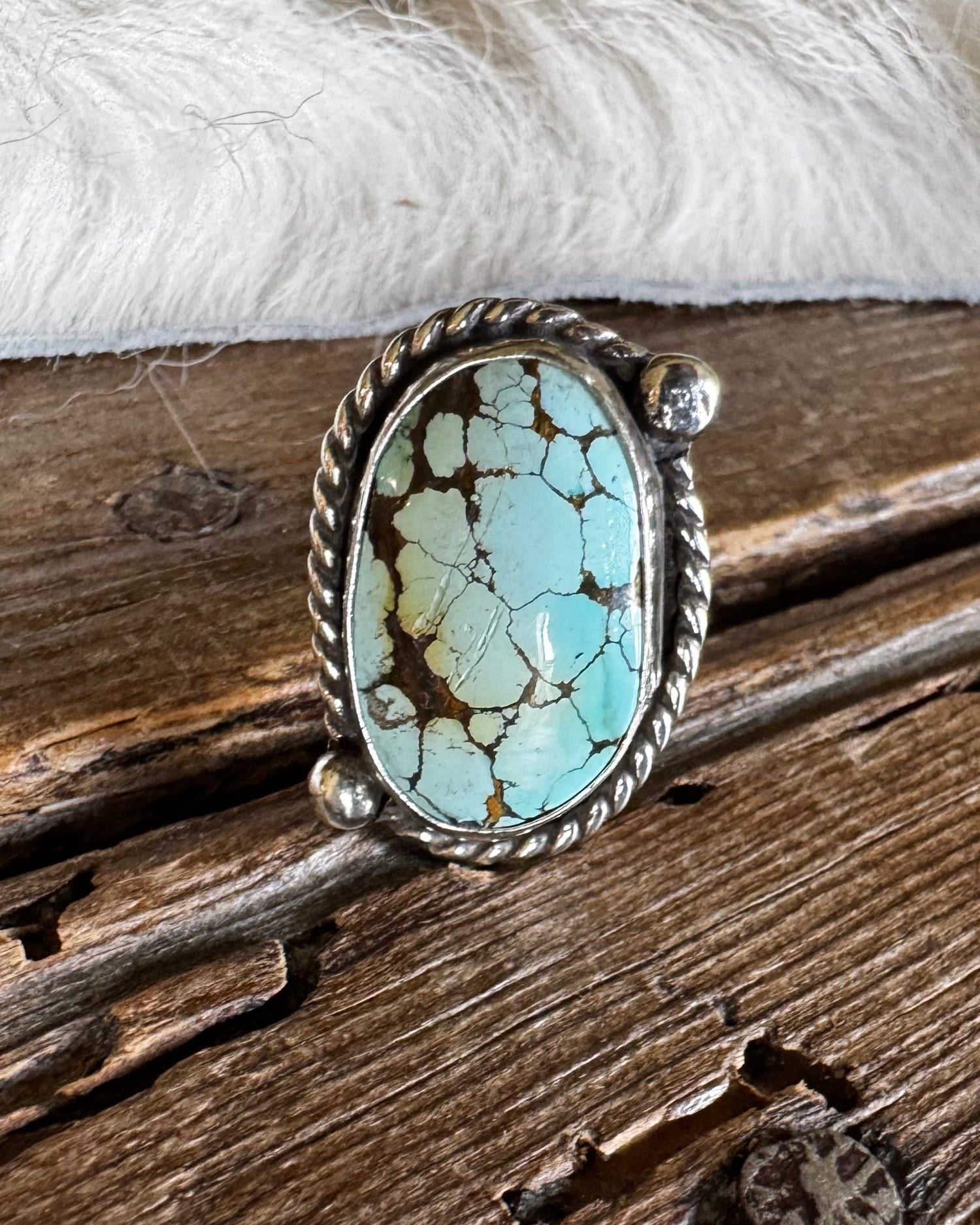 A silver ring featuring an oval turquoise stone with brown matrix veining, displayed on a rustic wooden surface with a white faux fur accent.