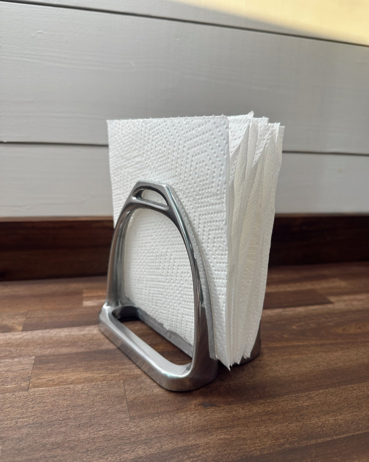 Metal stirrup-shaped napkin holder with white paper towels on a wooden surface.