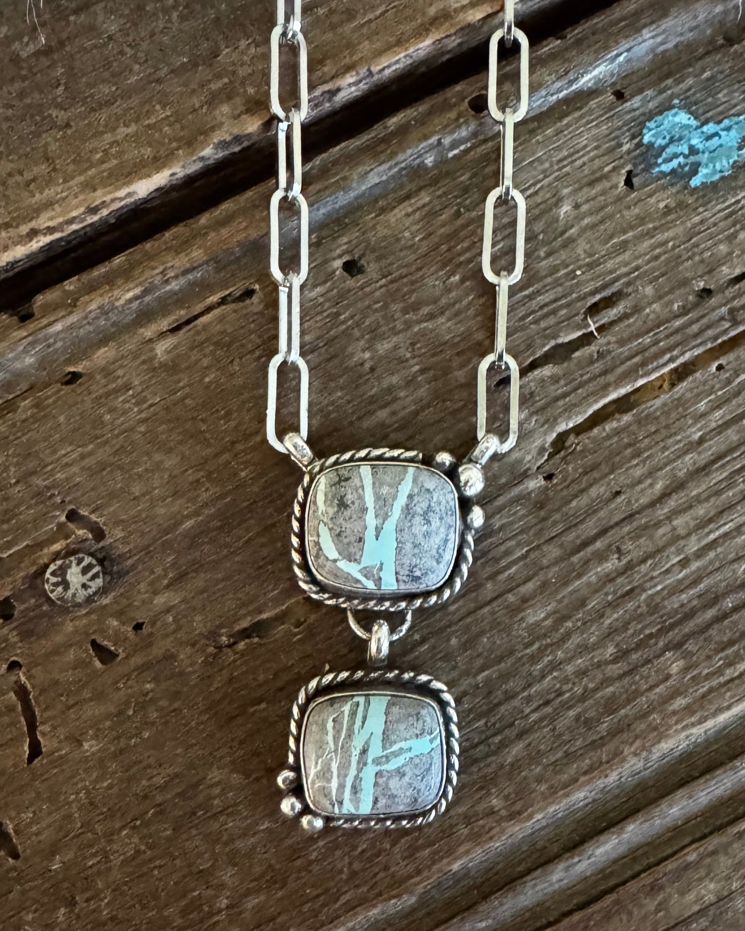 A silver link chain necklace with two stacked rectangular cabochon gemstone pendants, displayed on a rustic dark wood surface.
