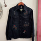 Black denim shirt with embroidered designs on a hanger against a white wall.