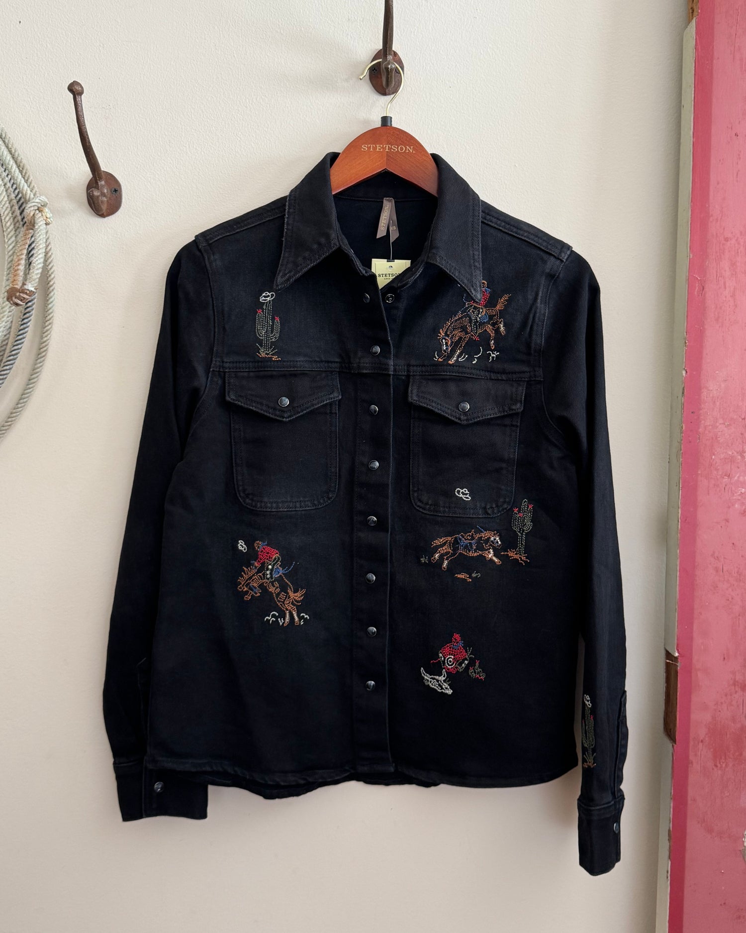 Black denim shirt with embroidered designs on a hanger against a white wall.