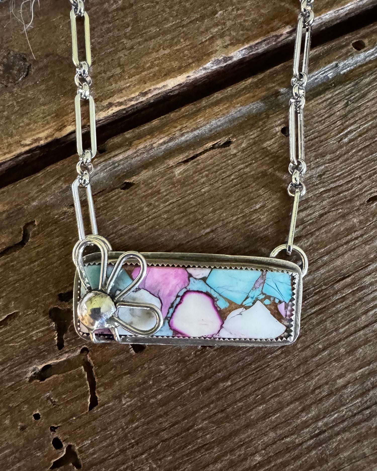 A rectangular pendant necklace with a colorful marbled inlay of turquoise, pink, and white, set in silver with a small butterfly charm, displayed on a rustic wooden surface.