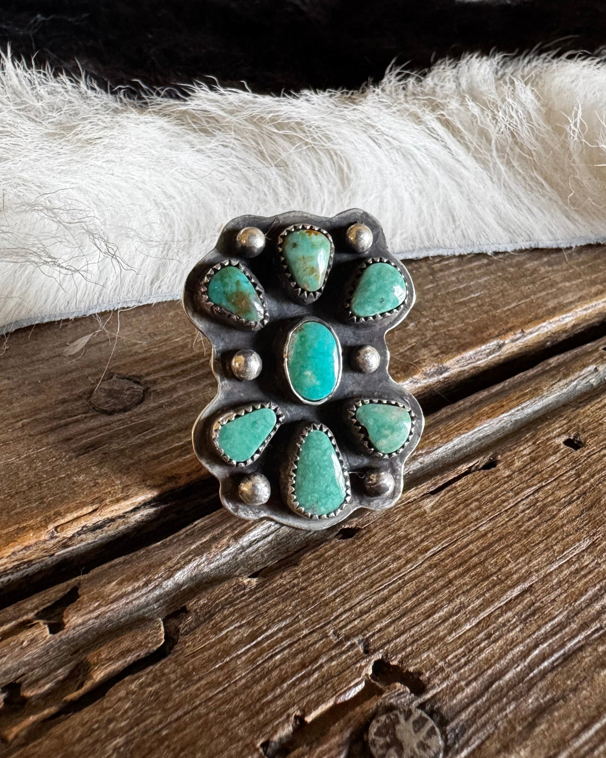 A silver ring with turquoise stones, displayed on a rustic wooden surface with a white fur trim and a blurred background.