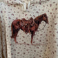 A cropped white hoodie with a small all-over floral print and a large graphic of a brown horse wearing a cowboy hat on the front, hanging on a wooden hanger.
