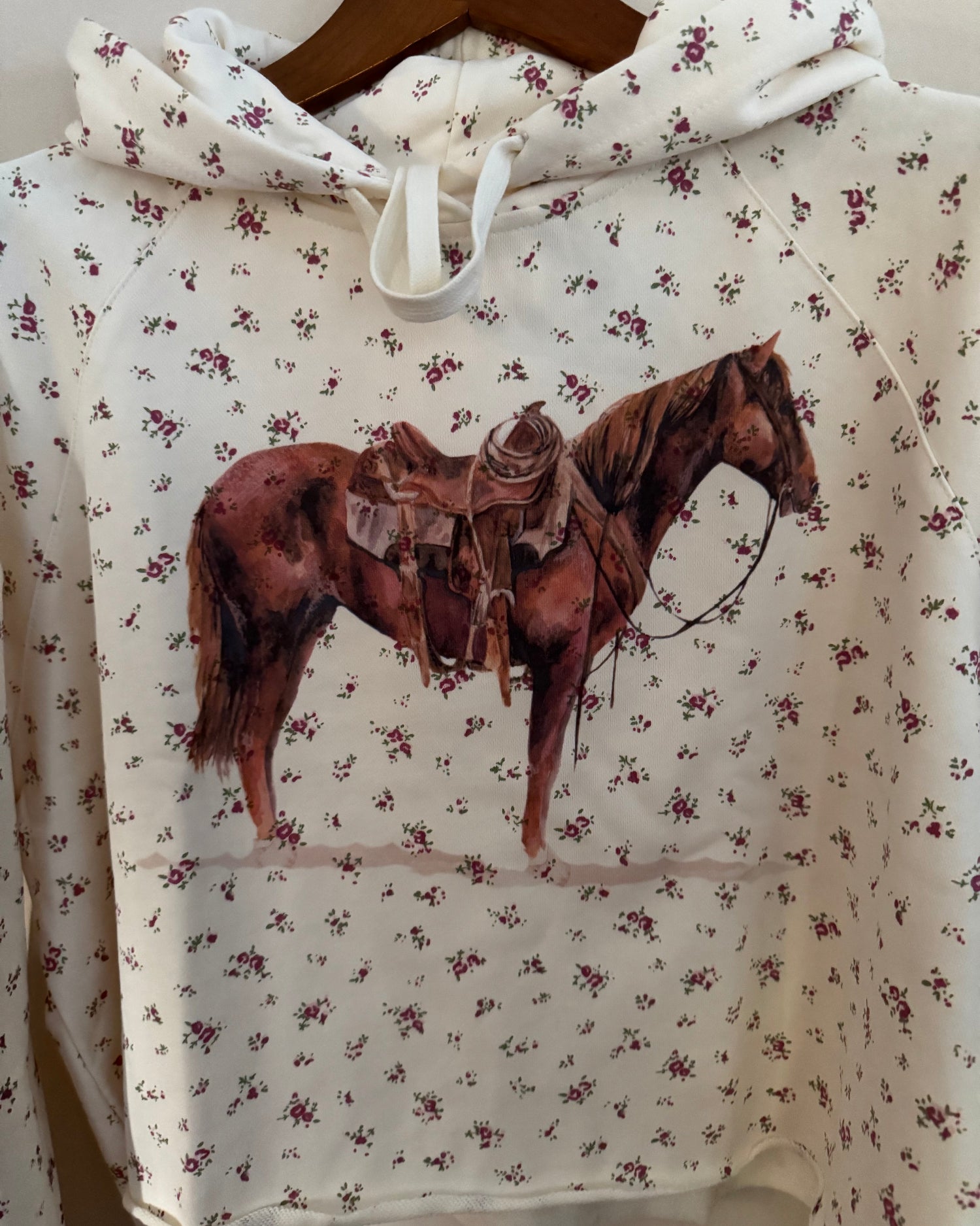 A cropped white hoodie with a small all-over floral print and a large graphic of a brown horse wearing a cowboy hat on the front, hanging on a wooden hanger.