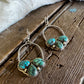 A pair of handmade silver hoop earrings with three green turquoise stones in a heart-shaped cluster setting, displayed on a rustic wooden surface next to a white faux fur trim.