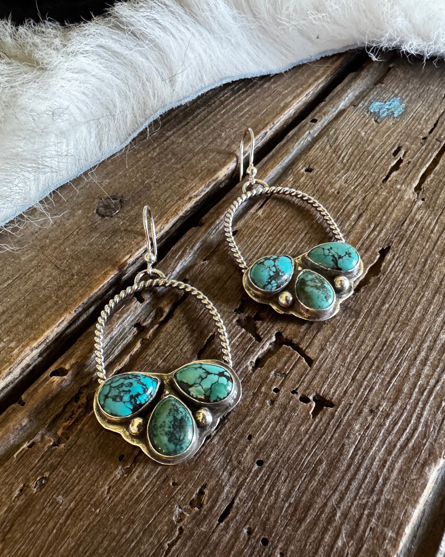 A pair of handmade silver hoop earrings with three green turquoise stones in a heart-shaped cluster setting, displayed on a rustic wooden surface next to a white faux fur trim.