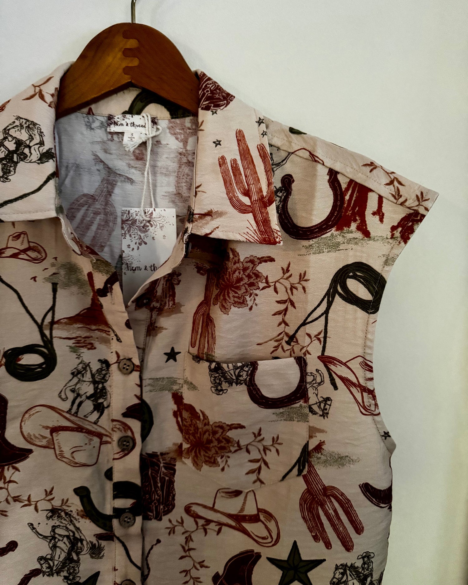 A sleeveless button-up shirt with a western-themed print featuring cacti, horseshoes, cowboy boots, stars, and riders on a beige background, hanging on a wooden hanger against a plain white wall.