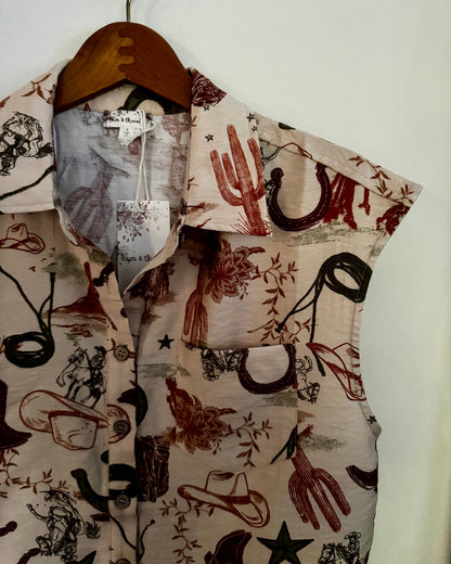 A sleeveless button-up shirt with a western-themed print featuring cacti, horseshoes, cowboy boots, stars, and riders on a beige background, hanging on a wooden hanger against a plain white wall.
