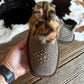 A person's hand holds a brown suede slipper with a leopard-print faux fur cuff and gold studded details, with another slipper in the background on a rustic wood floor and cowhide rug.