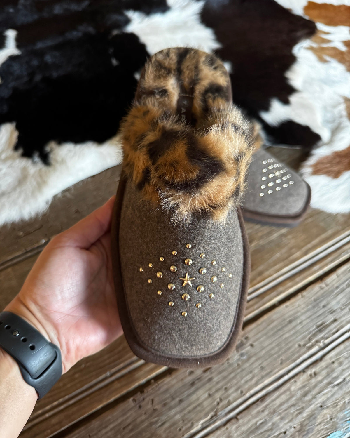 A person's hand holds a brown suede slipper with a leopard-print faux fur cuff and gold studded details, with another slipper in the background on a rustic wood floor and cowhide rug.