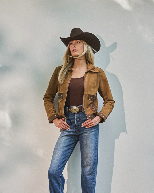 Woman wearing a brown jacket, jeans, and a cowboy hat against a light gray background