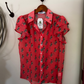 A coral pink short-sleeve button-up blouse with a green cactus print, hanging on a wooden hanger from a black wall hook.