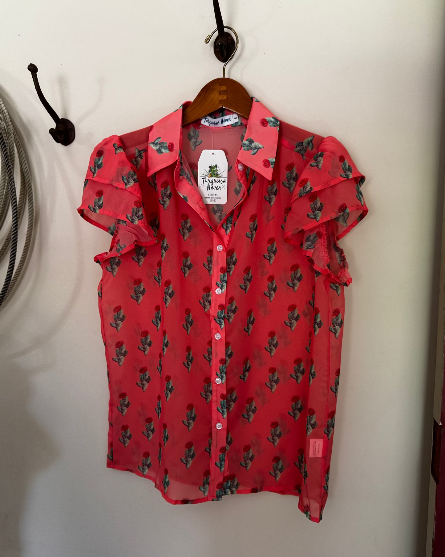 A coral pink short-sleeve button-up blouse with a green cactus print, hanging on a wooden hanger from a black wall hook.