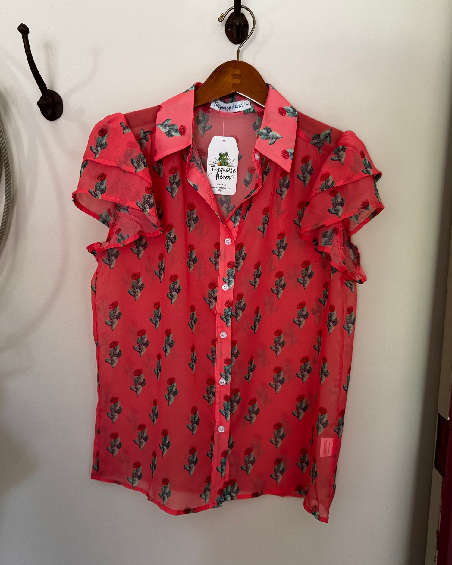 A red short-sleeve button-up blouse with a green cactus print, hanging on a wooden hanger against a white wall.