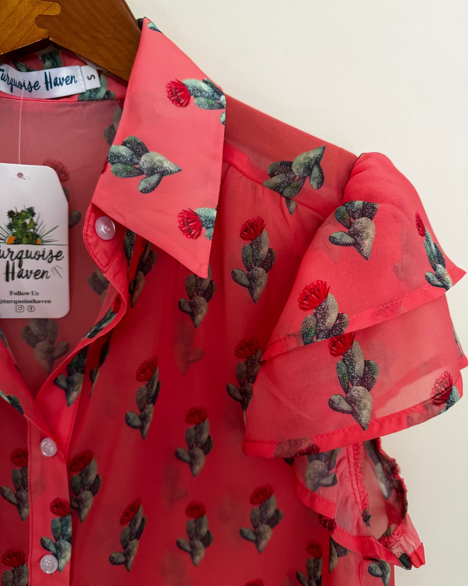 A coral pink sheer mesh blouse with a green and red cactus print, featuring ruffled sleeves and a collar, hanging on a wooden hanger with a 'Turquoise Haven' brand tag.