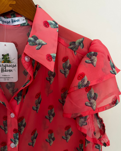 A coral pink sheer mesh blouse with a green and red cactus print, featuring ruffled sleeves and a collar, hanging on a wooden hanger with a 'Turquoise Haven' brand tag.